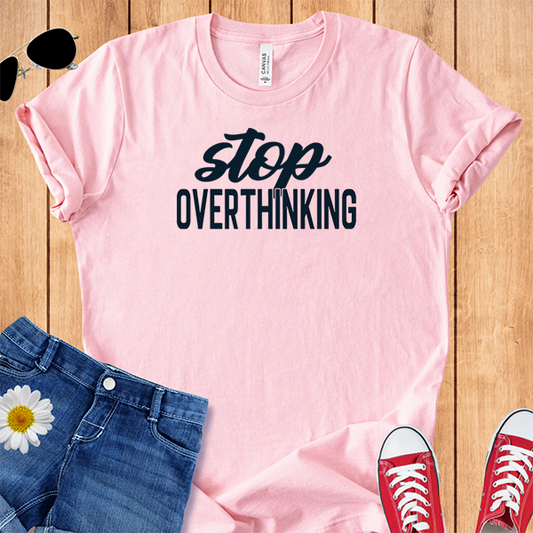 Pink t-shirt with 'stop overthinking' text, paired with jeans and sneakers on a wooden surface.