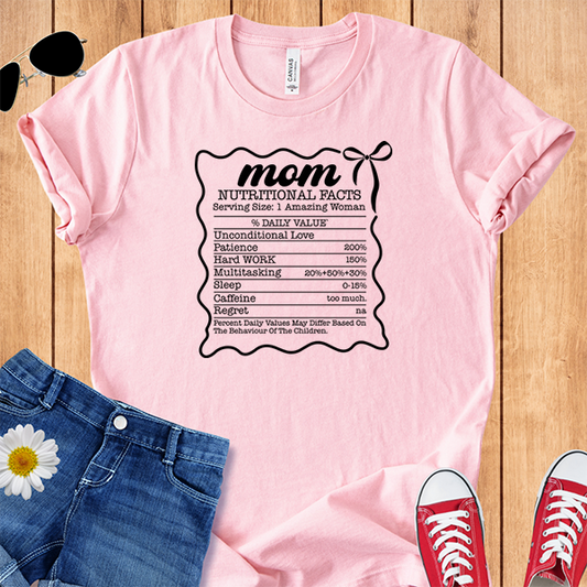 Pink t-shirt with 'mom nutritional facts' design on a wooden surface with jeans and sneakers.