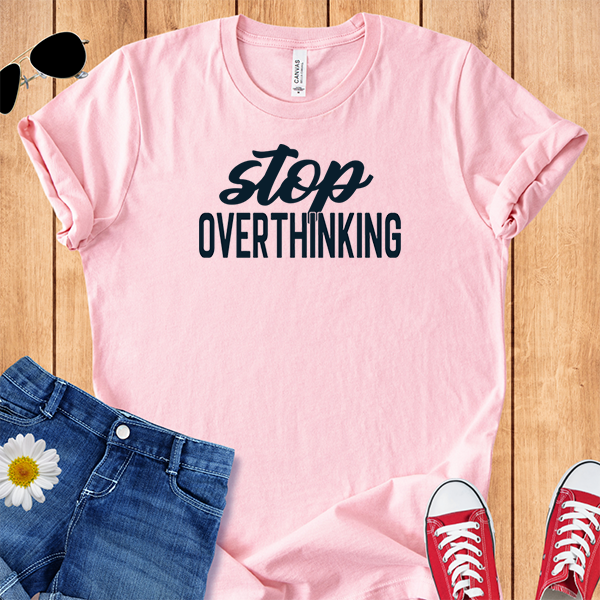 Pink t-shirt with 'stop overthinking' text, paired with jeans and sneakers on a wooden surface.